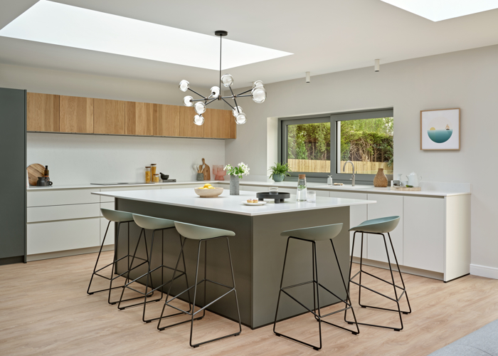 Wide shot of contemporary kitchen, with dark green kitchen island and tall cupboards, beige low cupboards and oak high cupboards, with glass pendant and modern stools