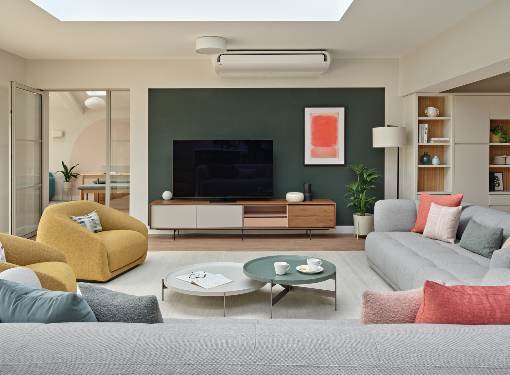 Sitting area within open plan space, with warm grey sofas, mustard coloured swivel armchairs, walnut tv unit and dark green wall colour accents of coral